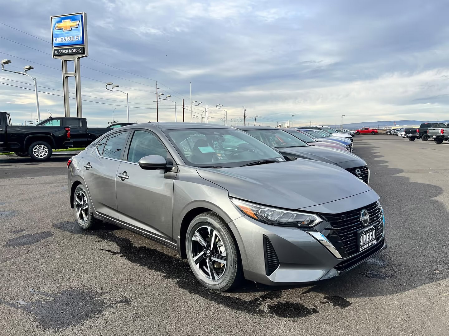 Gray Nissan sedan parked at Chevrolet dealership with cloudy sky and other vehicles in background