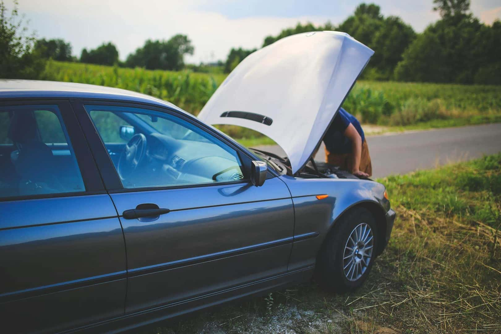 A person with a broken down car on the side of the road opening up their hood.