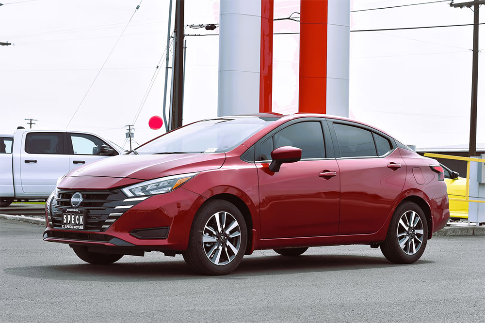 Deep red Nissan Versa parked in urban setting with power lines and white truck in background, sleek modern design